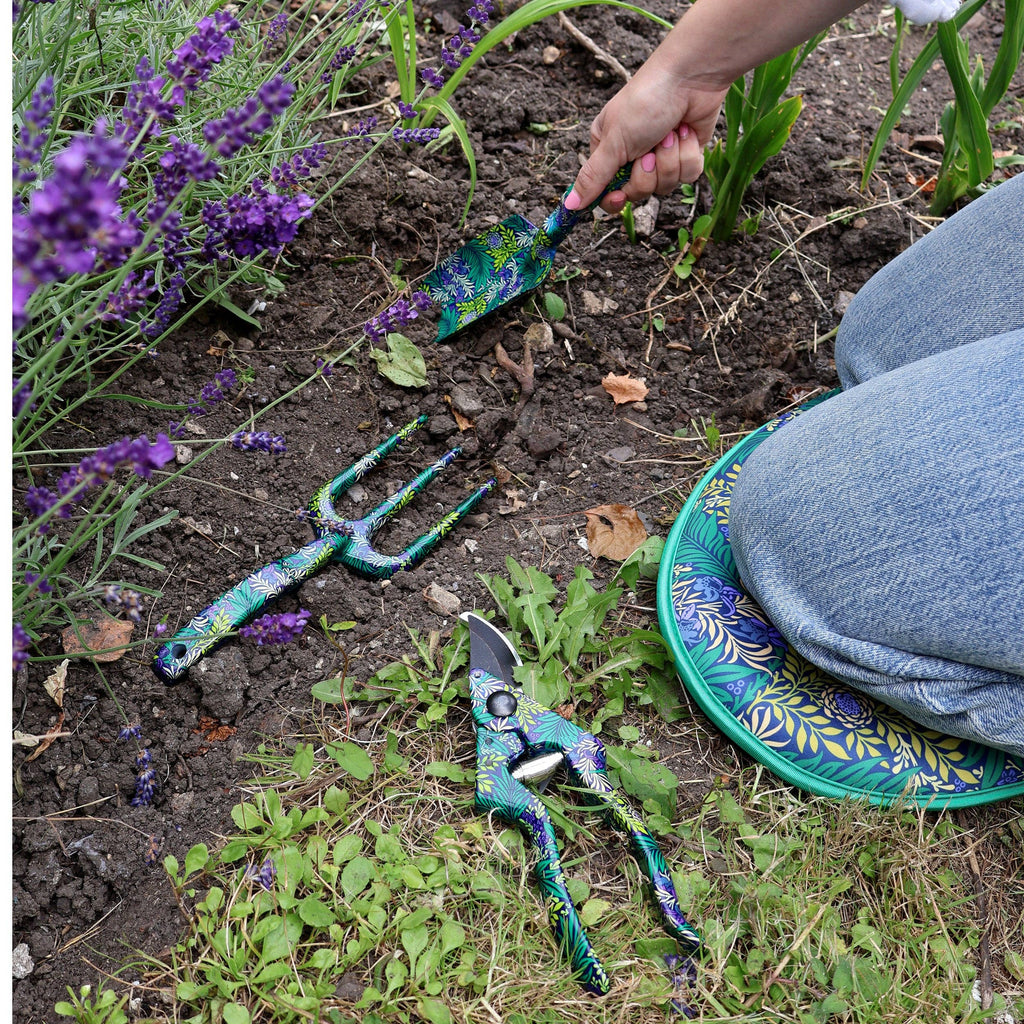 A person kneels on the ground, reaching for a pair of gardening tools beside them.