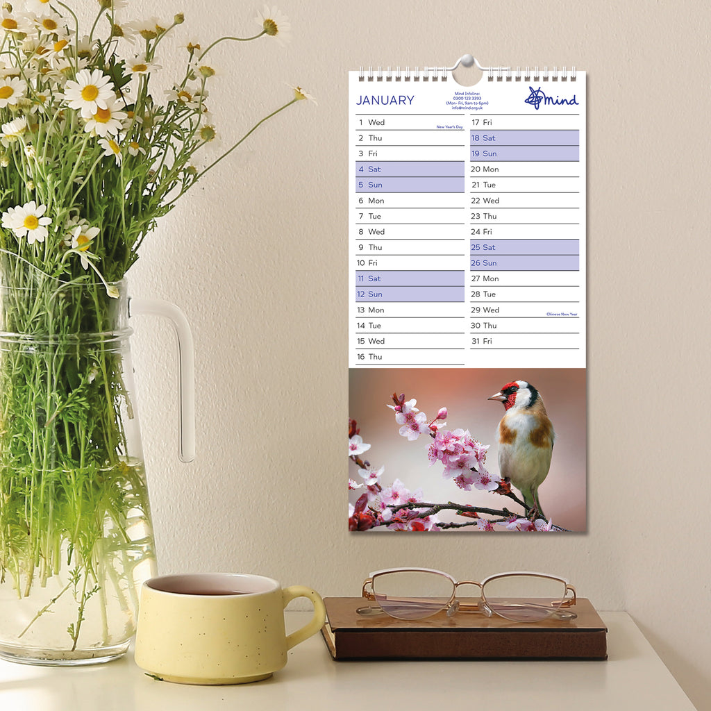 Inside the calendar on the month January with a bird perched on a branch with pink flowers hung on a wall in the background of a vase cup of coffee and glasses