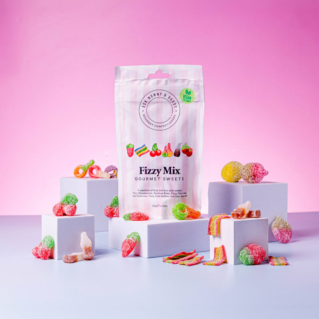 A colourful display of Fizzy Mix gourmet sweets in a pastel pink and white striped pouch, surrounded by an assortment of fizzy and sour jelly sweets shaped like strawberries, bottles, rings, and rainbows. The sweets are arranged on small white blocks against a vibrant pink gradient background, highlighting their bright colours and sugar-coated texture. The packaging features the text “Vegan Recipe” and illustrations of the sweets inside.