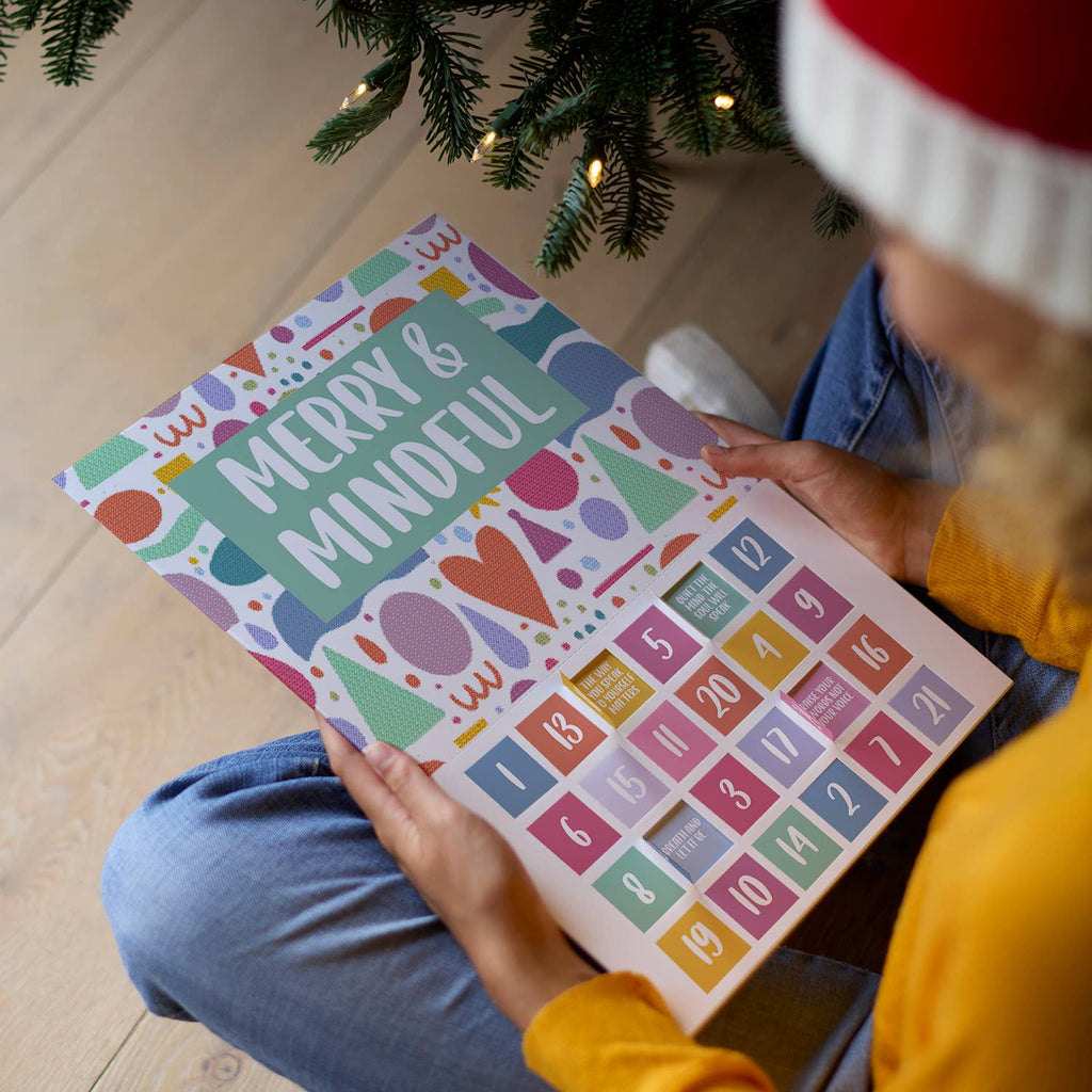 Festive and brightly coloured advent calendar with bold text saying "merry and mindful" on the front. A woman wearing a Santa hat is sitting cross-legged by the Christmas tree holding the advent calendar with both hands.