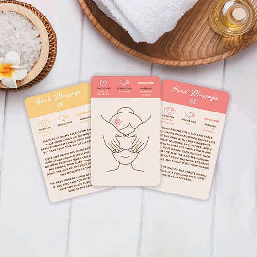 Three cards with a woman's picture diagram and massage techniques on a wooden surface surrounded by massage oils and salts.