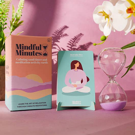 A box of mindfulness cards alongside a glass timer, encouraging calm and reflection on a studio pink backdrop.