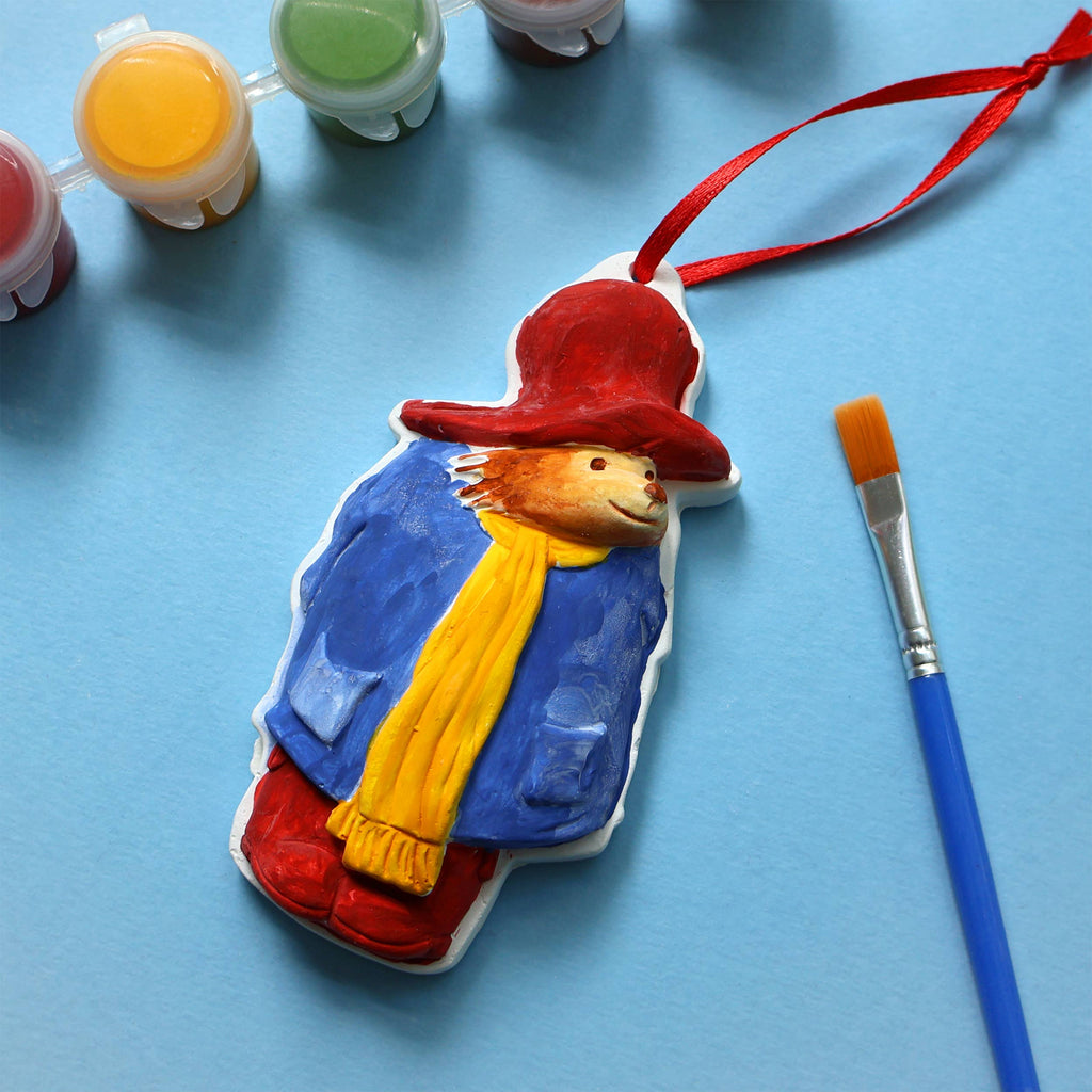 A small Paddington Bear figurine wearing a red hat and scarf, standing cheerfully with a friendly expression.