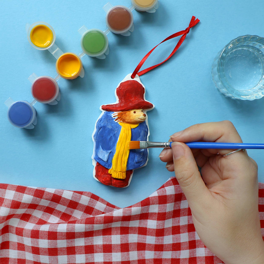 A person carefully paints a small Paddington decoration with colourful paint, focusing on details with a brush in hand.