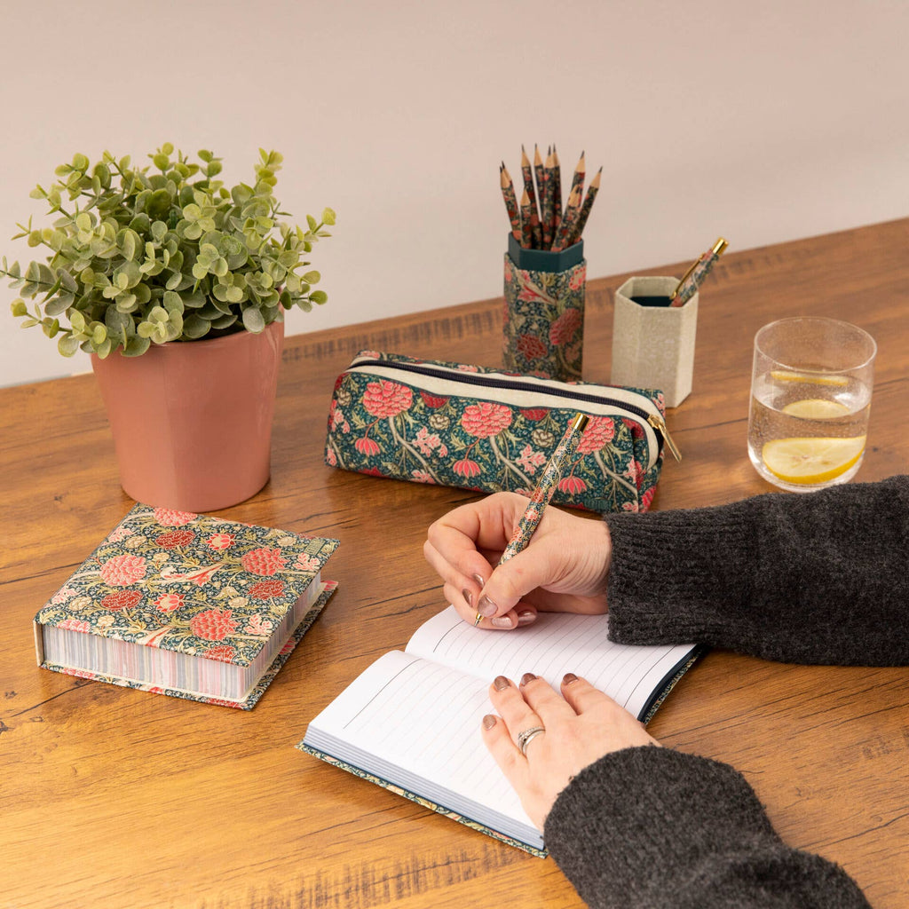 A lady's hand writing in a William Morris notebook, with parts of the collection visible, including a pencil set and pen set, available for sale on our website